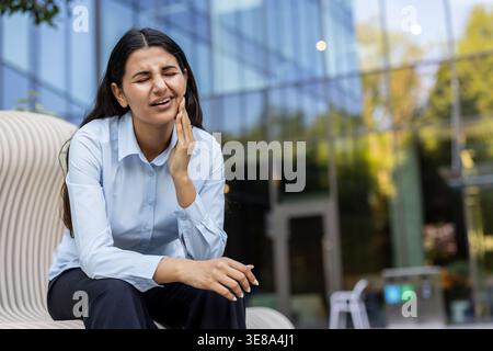 Jeune femme indienne grimaçant dans la douleur, tenant sa joue sur un banc de ville, souffrant de maux de dents intenses et d'inconfort à la mâchoire tout en étant habillée dans des affaires décontractées en plein air Banque D'Images