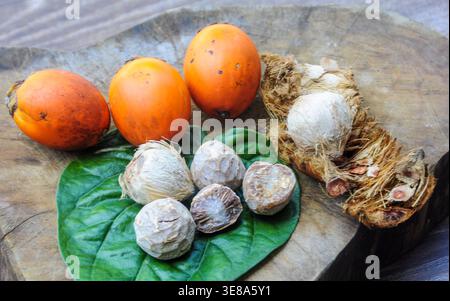 Assortiment de noix de bétel orange et vert avec coquilles de noix de bétel fissurées disposées sur une surface en bois altérée dans une composition de produit tropical rustique. Banque D'Images