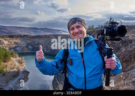 Souriant vidéaste masculin d'âge moyen donnant un pouce vers le haut tout en tenant une caméra à cardan sur le bord de la carrière de Racos avec le lac sarcelle visible dans la BA Banque D'Images