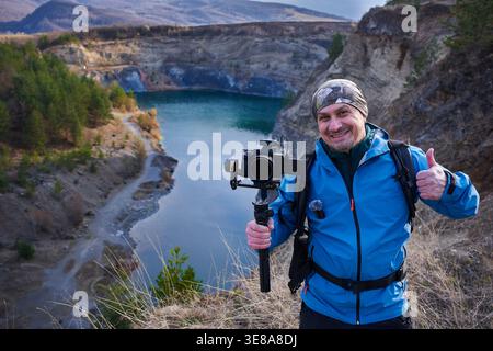Souriant vidéaste masculin d'âge moyen donnant un pouce vers le haut tout en tenant une caméra à cardan sur le bord de la carrière de Racos avec le lac sarcelle visible dans la BA Banque D'Images