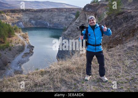 Souriant vidéaste masculin d'âge moyen donnant un pouce vers le haut tout en tenant une caméra à cardan sur le bord de la carrière de Racos avec le lac sarcelle visible dans la BA Banque D'Images