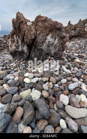 Une grande roche volcanique escarpée se trouve bien en évidence sur une plage couverte de galets lisses et variés, avec la mer visible au loin sous un ciel nuageux Banque D'Images