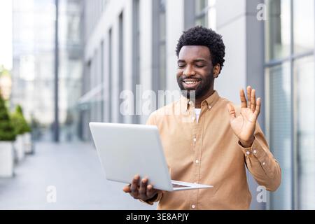 Homme afro-américain souriant et agitant devant un ordinateur portable pendant un appel vidéo extérieur, travaillant à distance depuis une rue urbaine avec des immeubles de bureaux modernes en arrière-plan Banque D'Images
