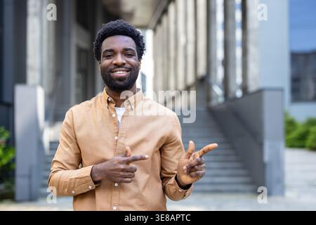 Homme souriant debout dehors devant un bâtiment moderne, pointant joyeusement ses doigts sur le côté, communiquant les options ou montrant la voie à suivre Banque D'Images