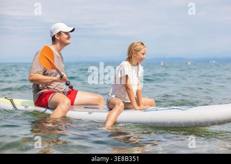 Père et fille profitent du temps sur un stand up paddleboard à la mer pendant l'été, partageant sourires et joie dans leur aventure aquatique. Banque D'Images