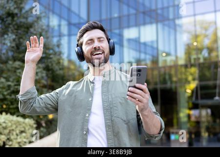 Homme enthousiaste avec des écouteurs fermant les yeux en profitant de la musique, chantant fort et tenant son smartphone tout en se tenant dehors devant un bâtiment moderne de la ville Banque D'Images