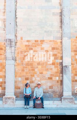 Pise, Italie : un couple assis ensemble contre un mur de pierre historique contrasté entre deux colonnes de marbre dans le centre-ville. Banque D'Images