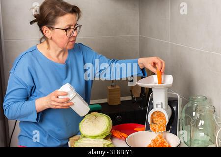Une femme travaille dans une cuisine pour préparer du chou fermenté. Elle utilise une râpe et mélange des carottes râpées tout en mesurant le sel. La cuisine a différents ustensiles et pots. Banque D'Images