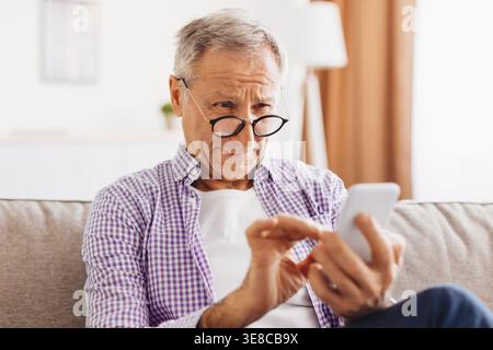 Homme senior regarde le téléphone tout en étant assis sur le canapé dans le salon pendant la journée Banque D'Images