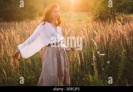 Jeune femme en blouse blanche et jupe beige marchant librement par prairie dorée profitant du coucher du soleil chaude lumière paisible atmosphère de la nature exprimant freedo Banque D'Images
