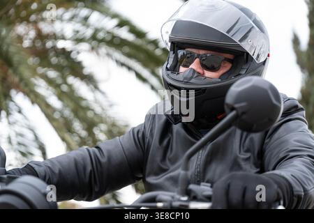 Homme senior actif sur une moto, portant une veste en cuir noir et un casque, conduisant à l'extérieur avec passion pour les voyages et la liberté sur route Banque D'Images
