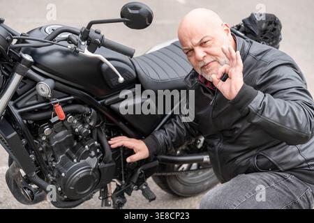 Homme chauve avec la barbe portant une veste en cuir noir, faisant un geste correct à côté de sa moto, montrant son approbation Banque D'Images
