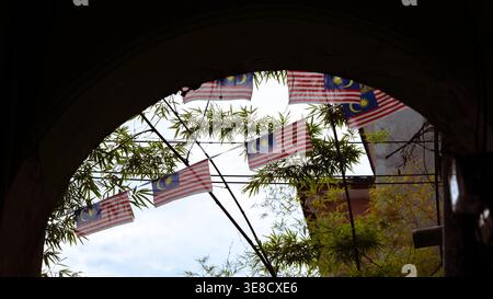Drapeaux nationaux malaisiens vus à travers une arche de pierre encadrée par des feuilles de bambou à George Town, Penang. Une composition patriotique et riche en patrimoine célébrant l'identité nationale au milieu du paysage de rue du patrimoine mondial de l'UNESCO. Banque D'Images