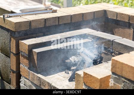 Barbecue en brique extérieur avec feu de charbon de bois brûlant et fumée montante, une installation de cuisine traditionnelle rustique dans un jardin. La lueur chaude des flammes évoque les rassemblements estivaux et le plaisir de cuisiner en plein air. Banque D'Images
