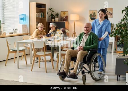 Homme âgé handicapé assis en fauteuil roulant tandis que jeune adulte femme aidant dans la maison de soins, soutenant le concept de soins aux personnes âgées. Utile pour les soins de santé, les soins infirmiers, le marketing de la vie assistée Banque D'Images