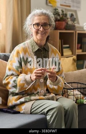 Portrait de femme senior caucasienne tricotant à la maison, souriant à la caméra et pratiquant un passe-temps créatif pour le concept de vieillissement actif. Utile pour le style de vie de retraite, le bien-être, le marketing des loisirs, vertical Banque D'Images