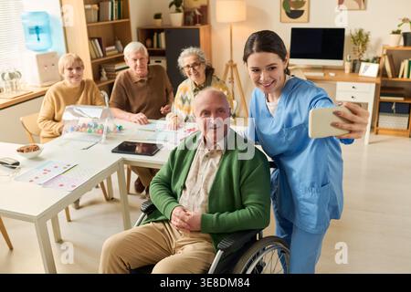 Jeune adulte femme soignante prenant selfie smartphone avec un homme âgé handicapé en fauteuil roulant pendant l'activité de groupe. Utile pour les soins aux aînés, maison de retraite, marketing de soins Banque D'Images
