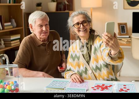 Couple caucasien senior jouant au bingo et prenant selfie smartphone à la maison, montrant une retraite active et une connexion numérique. Utile pour le mode de vie des aînés, le vieillissement, le marketing des soins sociaux Banque D'Images