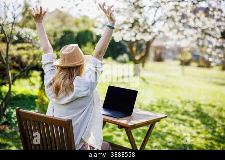 Femme étirant les bras tout en travaillant sur ordinateur portable dans le jardin de printemps. Soulagement des contraintes. Freelance prenant une pause pendant le travail administratif à distance Banque D'Images