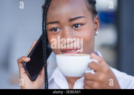 Gros plan d'une jeune femme noire parlant sur smartphone tout en tenant un café dans un café. Communication mobile conviviale, support client, entreprise à distance c Banque D'Images