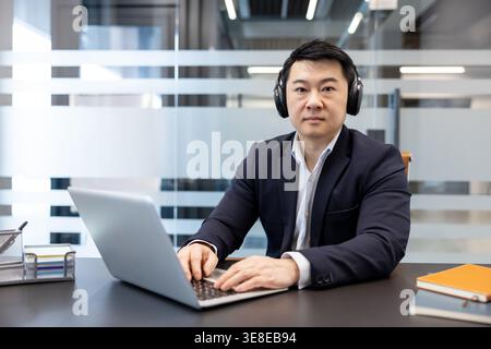 Homme d'affaires asiatique portant des écouteurs sans fil et tapant sur un clavier d'ordinateur portable, se concentrant sur le travail à un bureau dans un bureau contemporain de bureau flou Banque D'Images