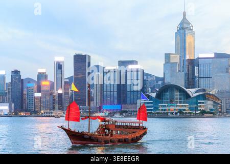 Jonque traditionnelle en bois avec des voiles rouges contre Hong Kong skyline pendant le coucher du soleil, scène urbaine emblématique Banque D'Images