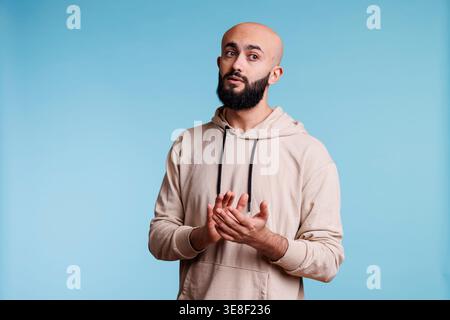 Jeune homme arabe applaudissant et soutenant la performance tout en détournant le regard. Personne barbu chauve dans des vêtements à capuche décontractés applaudissant avec les mains et montrant un geste de félicitations Banque D'Images