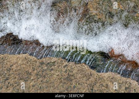 L'eau moussante coule rapidement sur la surface texturée des roches humides et des cailloux dans un ruisseau clair et peu profond, créant un motif abstrait dynamique de natu Banque D'Images