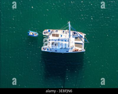 Vue aérienne d'un voilier catamaran blanc avec un petit canot bleu ancré dans une eau turquoise. Banque D'Images