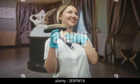 Femme dentiste en uniforme blanc tient le guide de teinte de dent et touche la joue avec la main gantée dans la construction de la clinique ; soins dentaires de confiance. Banque D'Images