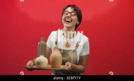 Femme tenant des vases de poterie sur un plateau en bois avec les deux mains, souriant et présentant la céramique en studio ; joie créativité. Banque D'Images