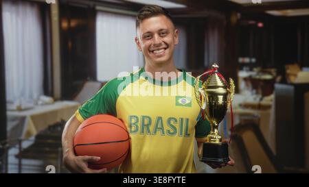 Homme tient le basket et le trophée dans un restaurant portant le maillot jaune du brésil et souriant avec la main visible sur le ballon ; fierté de célébration de la victoire. Banque D'Images
