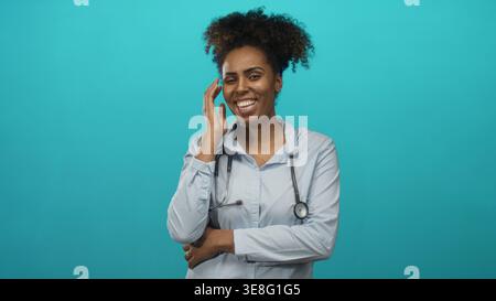 Jeune femme afro-américaine médecin portant un stéthoscope et une chemise bleue souriant largement en studio turquoise ; soin chaleur confiance. Banque D'Images
