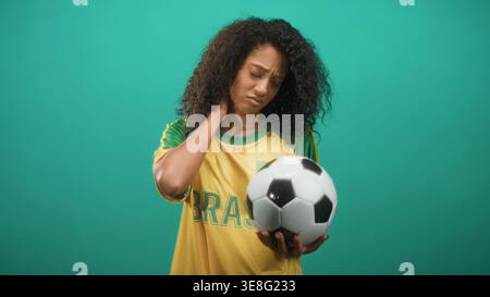 Adolescente tenant le ballon de football tient le cou en studio, portant le t-shirt de l'équipe du brésil et griffant avec les yeux fermés ; résilience de l'inconfort. Banque D'Images