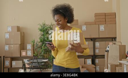 Femme tenant le colis et le smartphone, vérifiant l'étiquette d'expédition et le paquet dans un bâtiment de salle d'emballage ; esprit d'entreprise concentré. Banque D'Images