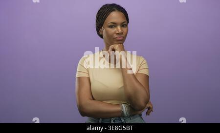 Femme avec la main sur le menton et les bras pliés debout dans le studio à côté du mur violet portant un tee-shirt beige et un Jean denim ; pensée contemplative. Banque D'Images