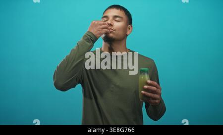 Jeune homme dégustant du jus vert d'une petite bouteille, main à bouche tout en tenant la bouteille sur fond de studio sarcelle ; bien-être calme. Banque D'Images