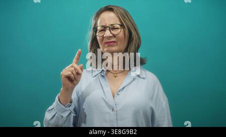 Femme senior pointe le doigt avec index relevé et main visible tout en portant des lunettes et une chemise bleu clair dans le studio ; avertissement de désapprobation. Banque D'Images