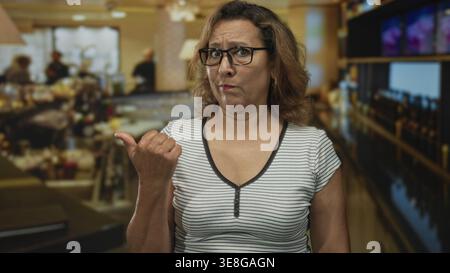 Une femme pointe le pouce sur le côté à l'intérieur d'un bâtiment de restaurant occupé près du comptoir et des étagères, portant des lunettes et un dessus rayé ; problème de doute. Banque D'Images