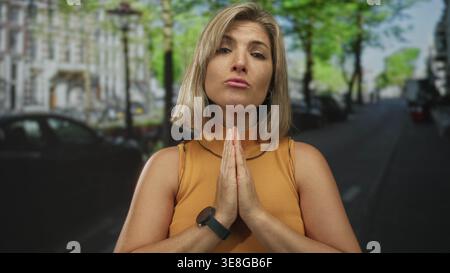 Jeune femme blonde avec les mains serrées à la poitrine se tient au milieu des voitures garées sur une rue urbaine ensoleillée animée ; sérénité. Banque D'Images
