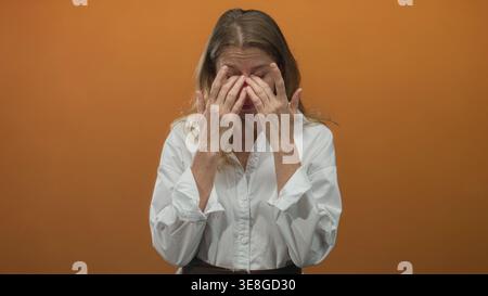 Femme pressant les mains aux yeux frottant le pont de nez dans le studio avec le mur orange portant une chemise blanche ; récupération de fatigue. Banque D'Images