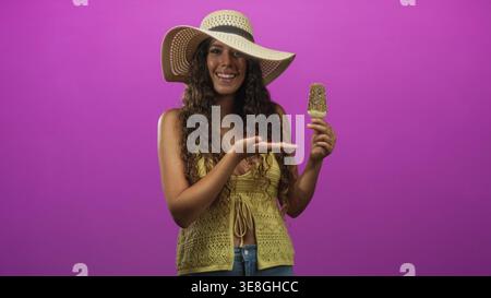 Femme hispanique portant un chapeau de soleil et un haut crochet jaune sourit, tient un popsicle et donne les pouces en studio ; joie d'été. Banque D'Images