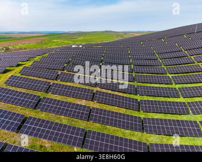 Des rangées de panneaux solaires couvrent un large champ sous le ciel bleu et le soleil. Des collines vertes s'étendent en arrière-plan, montrant une zone rurale. La configuration est utilisée pour garer Banque D'Images