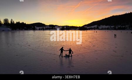 Vue aérienne de figures patinant sur le lac gelé avec une toile de fond de silhouettes d'arbres contre le ciel coloré du coucher de soleil, les Rousses, Bourgogne-Fran Banque D'Images