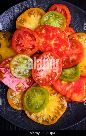 Tranches de tomates rouges, jaunes et vertes mélangées sur assiette foncée. Concept de salade fraîche de saison, produits biologiques naturels Banque D'Images