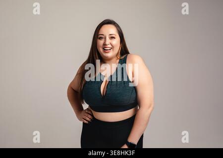 Concept de beauté inclusive. Souriant belle femme de taille plus avec silhouette athlétique courbée se tient en studio portant des vêtements de sport. Modèle féminin attrayant ex Banque D'Images