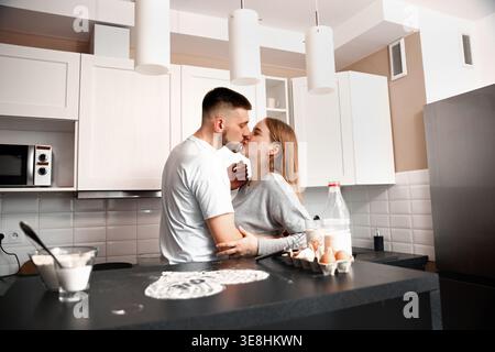 Un couple se tient dans une cuisine, échangeant un baiser pendant qu'ils préparent le petit déjeuner. Des ingrédients comme les œufs et la farine sont sur le comptoir, tandis que de la lumière se verse dans le Banque D'Images
