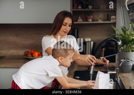Une mère aide son fils à se laver les mains à l'évier de la cuisine. Ils profitent tous les deux de ce temps ensemble pendant que la lumière entre par la fenêtre et t Banque D'Images