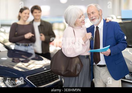 Jeune couple donne une voiture à leurs parents âgés. Un couple de personnes âgées heureux chez un concessionnaire automobile. Banque D'Images