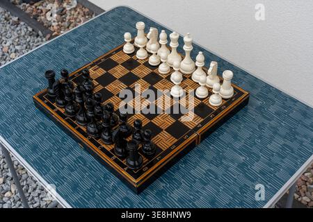 Échiquier orné avec des pièces noires et blanches sur une table pliante bleue à l'extérieur entourée de gravier et de mur blanc. Concept de jeu de plein air occasionnel Banque D'Images